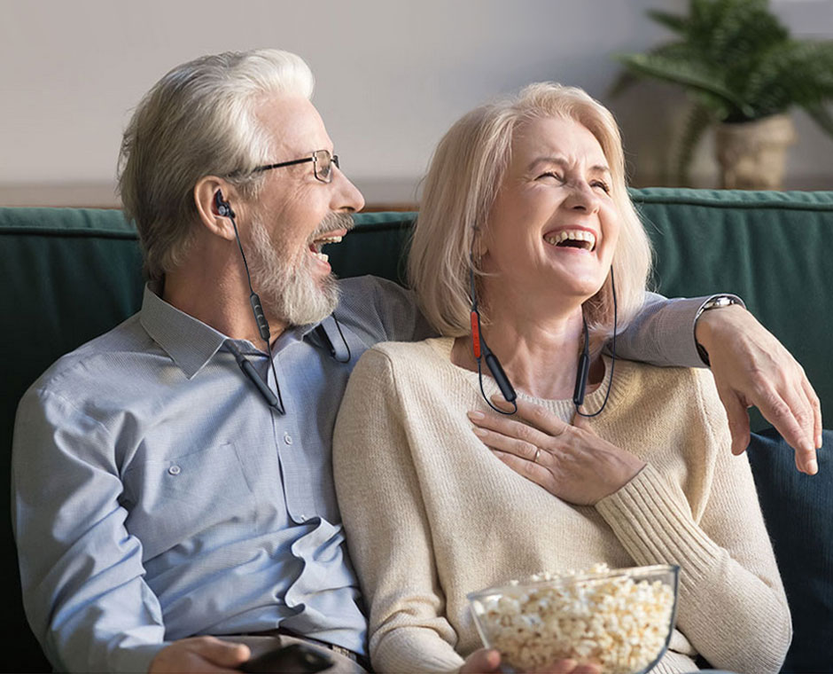 Older Couple laughing with nowl of popcorn and a remote in hand, each wearing a HT41866 Bluetooth in-ear headphone